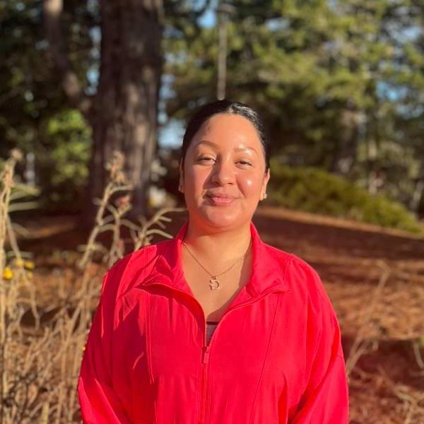 A headshot of Griselda. She smiles against a wooded background, wearing a bright red jacket.